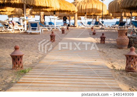 Wooden boardwalk leads across a sandy beach toward straw umbrellas sunbeds and the sea in a resort recreation area. Beach leisure vacation tourism resort lifestyle Wooden boardwalk leads across a sandy beach toward straw umbrellas sunbeds and the sea in a resort recreation area. Beach leisure vacation tourism resort lifestyle 140149309