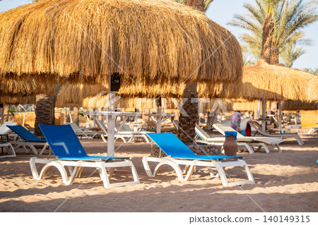 Empty blue sunbeds stand on a sandy beach beneath straw umbrellas with palm trees and resort facilities in the background. Beach leisure vacation tourism resort lifestyle Empty blue sunbeds stand on a sandy beach beneath straw umbrellas with palm trees and resort facilities in the background. Beach leisure vacation tourism resort lifestyle 140149315