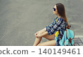 Stylish teenager girl with backpack, young woman sitting on city street on a summer day 140149461