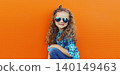 Happy cheerful kid girl with curly hair, joyful little child having fun in the city on orange wall 140149463