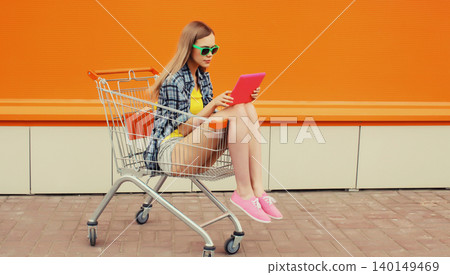 Modern young woman using digital tablet computer sitting in shopping trolley cart on city street 140149469