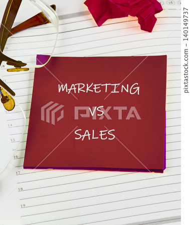 A vibrant workspace showcases a red sticky note with bold letters stating marketing versus sales, accompanied by glasses and delicate rose petals, symbolizing collaboration A vibrant workspace showcases a red sticky note with bold letters stating marketing versus sales, accompanied by glasses and delicate rose petals, symbolizing collaboration 140149737