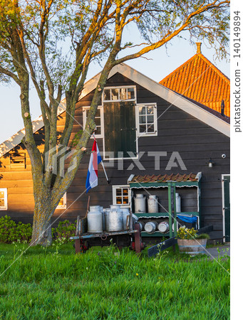 Traditional wooden Dutch farmhouse with national flag, green grass and warm sunset light, representing rural lifestyle, heritage architecture and countryside scenery in the Netherlands. 140149894