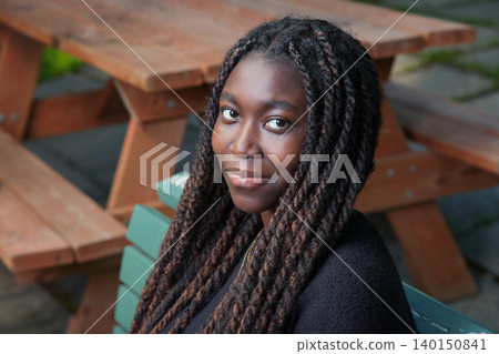 Portrait of a smiling young Black woman sitting on a green bench near a wooden picnic table Portrait of a smiling young Black woman sitting on a green bench near a wooden picnic table 140150841