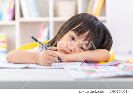 A tired little schoolgirl leaning her head on her arms on a desk while holding a pen during a boring study session. A tired little schoolgirl leaning her head on her arms on a desk while holding a pen during a boring study session. 140151014