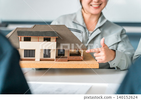 A woman in work clothes, working in the construction or building industry, uses a model house to conduct business negotiations, explanations, and presentations. A woman in work clothes, working in the construction or building industry, uses a model house to conduct business negotiations, explanations, and presentations. 140152624