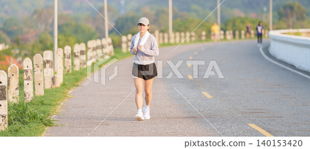 Active Asian woman jogging with a towel around her neck in the park at morning. Female runner exercising in summer for healthy lifestyle and fitness workout outdoors. Wellness and sport concepts Active Asian woman jogging with a towel around her neck in the park at morning. Female runner exercising in summer for healthy lifestyle and fitness workout outdoors. Wellness and sport concepts 140153420
