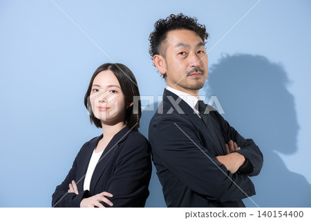 A businessman and a woman in suits stand back-to-back with their arms crossed, looking directly at the camera, their expressions serious and earnest, conveying leadership and trust. A businessman and a woman in suits stand back-to-back with their arms crossed, looking directly at the camera, their expressions serious and earnest, conveying leadership and trust. 140154400