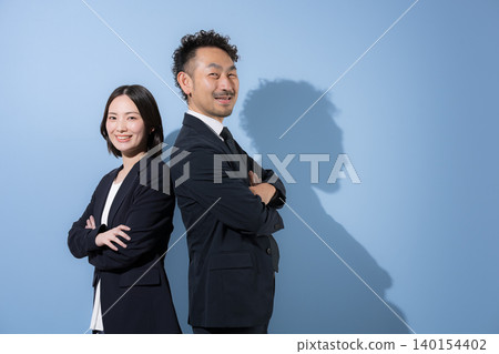 A businessman and his wife in suits stand back-to-back with their arms crossed, looking directly at the camera; their smiles convey leadership and a B2B image. A businessman and his wife in suits stand back-to-back with their arms crossed, looking directly at the camera; their smiles convey leadership and a B2B image. 140154402