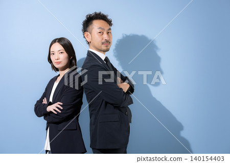 A businessman and a woman in suits stand back-to-back with their arms crossed, looking directly at the camera, their expressions serious and earnest, conveying leadership and trust. A businessman and a woman in suits stand back-to-back with their arms crossed, looking directly at the camera, their expressions serious and earnest, conveying leadership and trust. 140154403