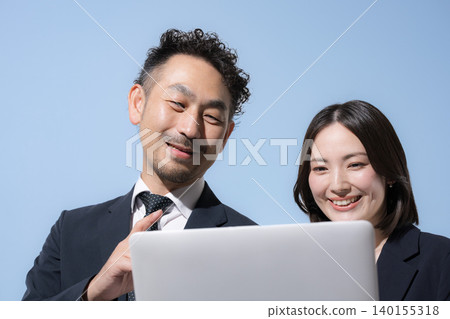 A close-up of a man and woman in business suits looking at a computer together against a light blue background. A close-up of a man and woman in business suits looking at a computer together against a light blue background. 140155318