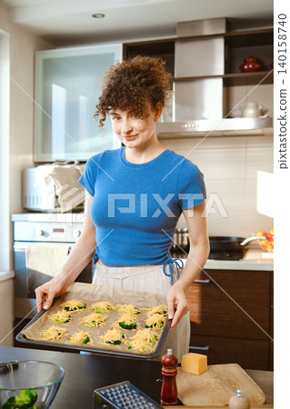 Cheerful woman holds a baking tray with smashed broccoli covered with grated cheese Cheerful woman holds a baking tray with smashed broccoli covered with grated cheese 140158740