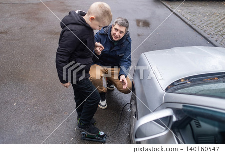 Father and son pumping a car tire with a foot pump on a city street. Caucasian man teaches his boy how to inflate automobile wheels, lifestyle family outdoor activity. Father and son pumping a car tire with a foot pump on a city street. Caucasian man teaches his boy how to inflate automobile wheels, lifestyle family outdoor activity. 140160547