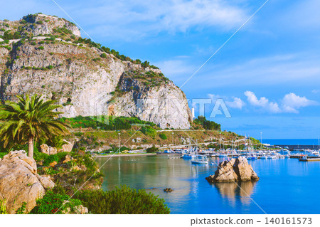 A picturesque coastal landscape featuring a rugged cliffside overlooking a tranquil bay dotted with numerous yachts and sailboats docked at a marina A picturesque coastal landscape featuring a rugged cliffside overlooking a tranquil bay dotted with numerous yachts and sailboats docked at a marina 140161573