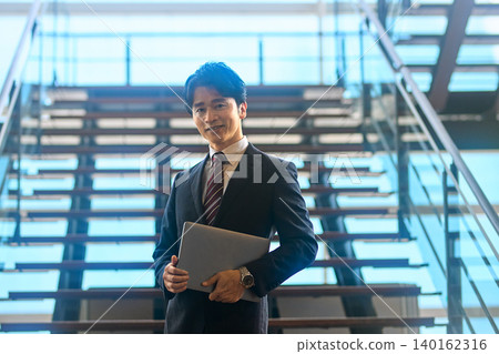 一位商務人士站在樓梯前,手裡拿著一台筆記型電腦。 一位商務人士站在樓梯前,手裡拿著一台筆記型電腦。 140162316