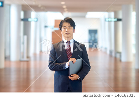 一位商務人士站在寬敞的走道裡,手持筆記型電腦,拍攝全身照。 一位商務人士站在寬敞的走道裡,手持筆記型電腦,拍攝全身照。 140162323