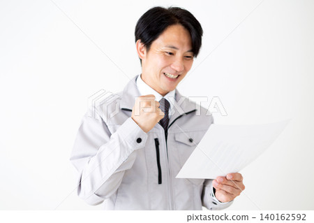 A middle-aged businessman in gray work clothes makes a triumphant gesture while looking at documents against a white background. A middle-aged businessman in gray work clothes makes a triumphant gesture while looking at documents against a white background. 140162592