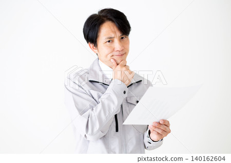 一位身穿灰色工作服的中年商人,在白色背景前看著文件,若有所思。 一位身穿灰色工作服的中年商人,在白色背景前看著文件,若有所思。 140162604