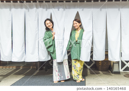 身穿浴衣的成年女性從溫泉旅館的白色簾子後面探出頭來，臉上帶著微笑。 140165468