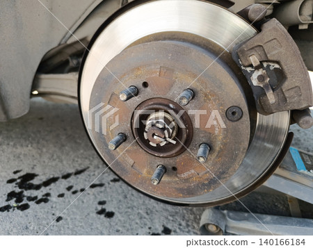 Close-up shot shows a car's disc brake assembly during maintenance. The wheel is removed, exposing a rusty wheel hub with studs, a shiny brake rotor, a caliper, and a jack lifting the vehicle Close-up shot shows a car's disc brake assembly during maintenance. The wheel is removed, exposing a rusty wheel hub with studs, a shiny brake rotor, a caliper, and a jack lifting the vehicle 140166184