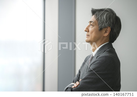 A senior businessman, arms crossed, looks out the window (profile view). A senior businessman, arms crossed, looks out the window (profile view). 140167751