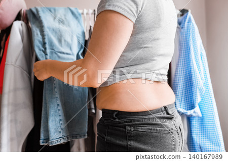 Overweight woman facing a wardrobe problem and clothing annoyance from weight gain. She holds jeans, her belly showing in tight pants Overweight woman facing a wardrobe problem and clothing annoyance from weight gain. She holds jeans, her belly showing in tight pants 140167989
