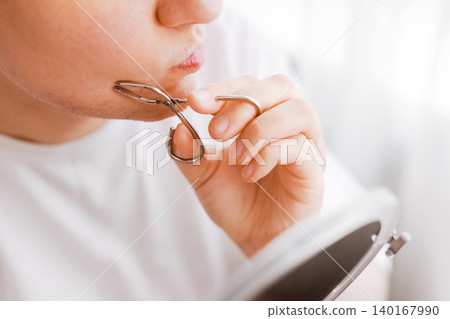 Woman plucks unwanted facial hair from her chin using tweezers by a mirror. A home beauty routine for hormonal hair removal and self-care Woman plucks unwanted facial hair from her chin using tweezers by a mirror. A home beauty routine for hormonal hair removal and self-care 140167990