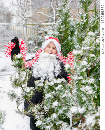 Child dressed as Santa with white beard and red tinsel garland standing outdoors Child dressed as Santa with white beard and red tinsel garland standing outdoors 140169882