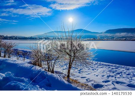 Early morning in winter at Lake Yamanaka: The frozen lake surface and frost-covered trees sparkle in the morning sun, creating a snowy landscape. Early morning in winter at Lake Yamanaka: The frozen lake surface and frost-covered trees sparkle in the morning sun, creating a snowy landscape. 140170530