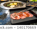 Slices of raw marinated beef in a black tray with metal tongs. Korean BBQ or Japanese yakiniku restaurant scene with a charcoal grill in the background. Fresh meat prepared for grilling. 140170669