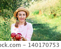 Portrait of woman in hat offering fresh red apples in orchard during warm sunny autumn day outdoors 140170753