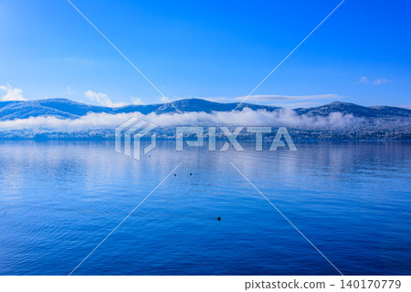 A minimalist background image of a winter lakeside enveloped in silence, with a clear blue sky and snow-covered trees. 140170779