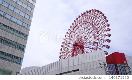 Modern Ferris wheel structure with a tall glass building against a cloudy sky Modern Ferris wheel structure with a tall glass building against a cloudy sky 140171930