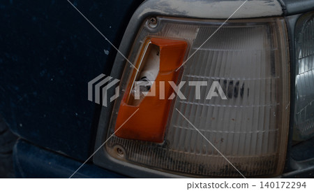 Close up view of a classic car showing a broken orange turn signal indicator lens next to the main headlight housing. The damaged plastic assembly highlights an auto repair need. Close up view of a classic car showing a broken orange turn signal indicator lens next to the main headlight housing. The damaged plastic assembly highlights an auto repair need. 140172294
