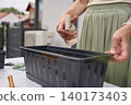 Woman waters soil in planter box with spray bottle on outdoor gardening table 140173403