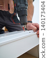 Man driving screw into drawer panel with cordless drill, hands close to the furniture part 140173404