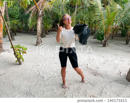 Young happy woman volunteer holding a black bag and showing a thumbs up while standing in a tropical environment Young happy woman volunteer holding a black bag and showing a thumbs up while standing in a tropical environment 140173815
