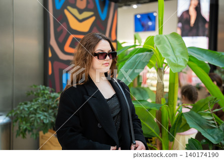 Young Caucasian woman wearing sunglasses stands near tropical plants inside a modern shopping mall with stores and decorative elements. Retail lifestyle and fashion scene 140174190