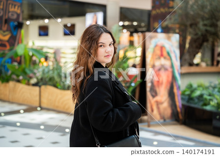 Young Caucasian woman with a black shoulder bag looks toward the camera while standing in a modern shopping mall with plants and stores. Retail lifestyle and consumer scene Young Caucasian woman with a black shoulder bag looks toward the camera while standing in a modern shopping mall with plants and stores. Retail lifestyle and consumer scene 140174191