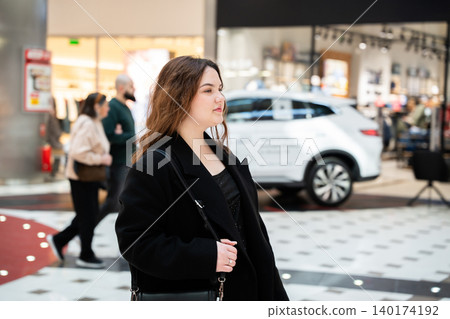 Young Caucasian woman with a shoulder bag walks through a modern shopping mall with stores visitors and a vehicle display in the background. Retail lifestyle and consumer scene 140174192