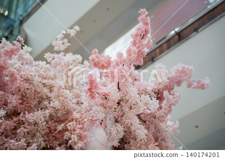 Pink blossom branches form a decorative floral installation inside a shopping mall with dense clusters of flowers and soft pastel tones. Retail decoration and lifestyle scene Pink blossom branches form a decorative floral installation inside a shopping mall with dense clusters of flowers and soft pastel tones. Retail decoration and lifestyle scene 140174201