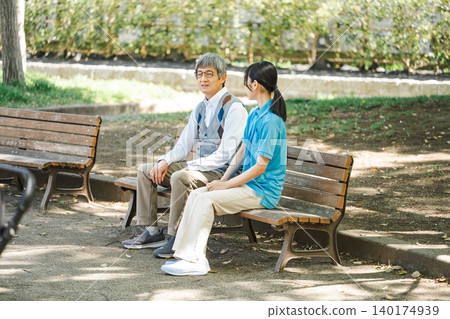 A caregiver and a senior male client sitting and talking on a park bench. A caregiver and a senior male client sitting and talking on a park bench. 140174939