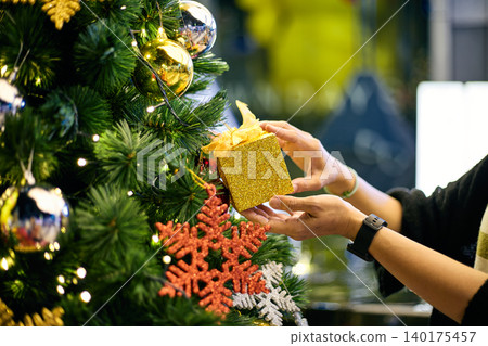 Hands decorating Christmas tree with glittering golden gift box among festive ornaments and warm fairy lights, capturing holiday celebration spirit, seasonal decoration tradition, joyful Christmas Hands decorating Christmas tree with glittering golden gift box among festive ornaments and warm fairy lights, capturing holiday celebration spirit, seasonal decoration tradition, joyful Christmas 140175457