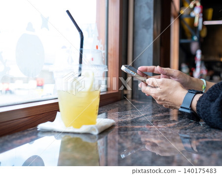 Person using smartphone beside refreshing iced drink at cafe window seat, representing mobile lifestyle, social media browsing, digital communication, remote work and modern relaxation concept Person using smartphone beside refreshing iced drink at cafe window seat, representing mobile lifestyle, social media browsing, digital communication, remote work and modern relaxation concept 140175483