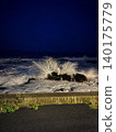 The waves crash against the rocky reefs at night, creating spray and a rough sea. 140175779