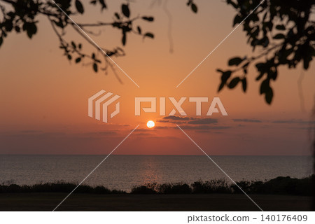 In the foreground, the silhouettes of land and vegetation are visible against an orange sunset sky, with the sun and the clearly defined horizon of the sea. In the foreground, the silhouettes of land and vegetation are visible against an orange sunset sky, with the sun and the clearly defined horizon of the sea. 140176409