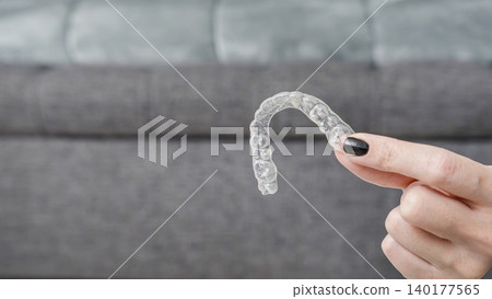 Transparent dental aligner held by a teenager with black nail polish, sitting on a gray couch in a modern living room setting Transparent dental aligner held by a teenager with black nail polish, sitting on a gray couch in a modern living room setting 140177565