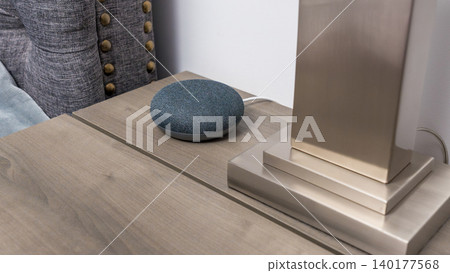 Smart home speaker on wooden nightstand beside a modern lamp with a gray upholstered headboard in a contemporary bedroom setting Smart home speaker on wooden nightstand beside a modern lamp with a gray upholstered headboard in a contemporary bedroom setting 140177568