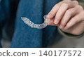 Person holding clear dental aligner with hand, wearing casual clothing, seated on a floor with denim jeans and a soft surface in the background 140177572