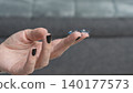Close-up of a hand holding two contact lenses with dark polished nails against a blurred background of a gray couch 140177573